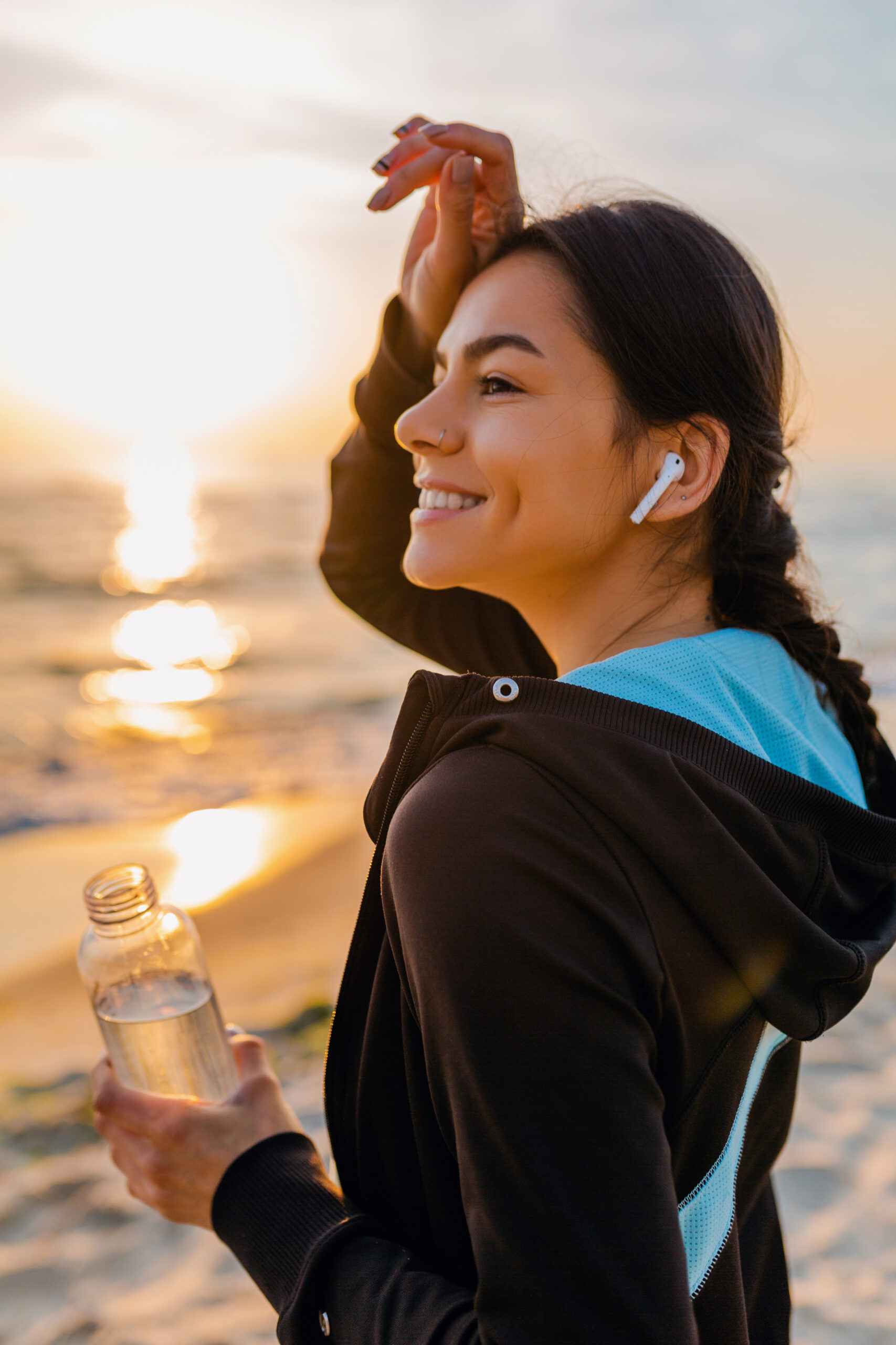 attractive slim woman doing sport exercises on morning sunrise beach in sports wear, thirsty drinking water in bottle, healthy lifestyle, listening to music on wireless earphones, hot summer day
